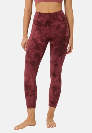 Vrouw draagt hooggetailleerde, bordeaux tie-dye legging en bijpassende sportbeha, staand op blote voeten tegen een effen achtergrond.