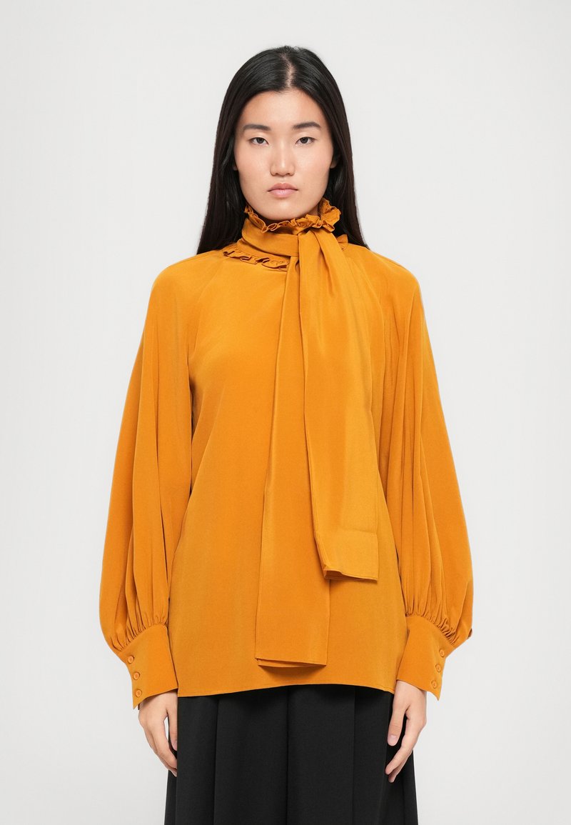 Mosterdgele blouse met lange, opgeblazen mouwen, gekruide kraag en strikdetail. Gemaakt van gladde stof met knoopmanchetten.