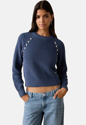 Modro pleten pulover z rebrasto teksturo, okroglim izrezom in okrasnimi belimi gumbi ob ramenih; skrajšan kroj. Nošen z modrimi kavbojkami.