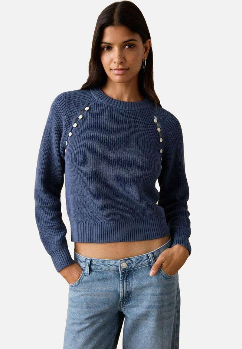 Plavi pleteni džemper s rebrastom teksturom, okruglim izrezom i ukrasnim bijelim gumbima duž ramena; kratkog kroja. Nošen uz plave traperice.