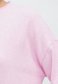 Ροζ ανοιχτόπλεκτο πουλόβερ με κοντινή λήψη της υφής του υφάσματος, που διαθέτει στρογγυλή λαιμόκοψη και ραφές με πέταγμα στους ώμους.