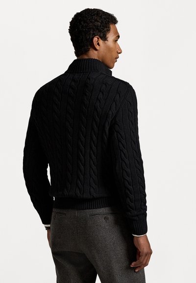 Maglione nero a maglia con colletto alto e orlo a coste, caratterizzato da motivi testurizzati. Il modello indossa pantaloni grigi, visto da dietro.