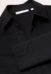 Chemise noire à col classique, patte de boutonnage et poignets repliés. Texture lisse et détail de surpiqûres subtils partout.