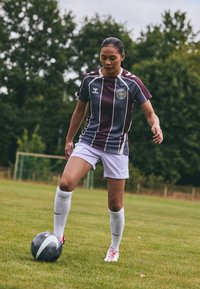 Athlète portant un maillot rayé bordeaux et bleu, un short blanc et des chaussettes blanches jusqu'aux genoux, dribbler un ballon de football noir et blanc sur l'herbe.