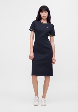 Donna che indossa un vestito aderente blu navy fino al ginocchio con rifiniture gialle su maniche e colletto, in piedi davanti a uno sfondo chiaro e semplice.