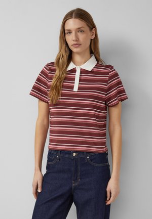 Frau mit langen blonden Haaren, die ein kurzärmeliges, gestreiftes Poloshirt in Weinrot, Pink und Weiß sowie dunkelblaue Jeans trägt, vor einem grauen Hintergrund.