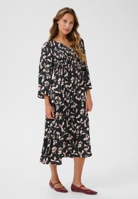 Rochie florală neagră cu bust plisat, mâneci lungi tip evazat și fustă de lungime medie. Prezintă modele florale în nuanțe de roz și crem. Pantofi plata roșii.