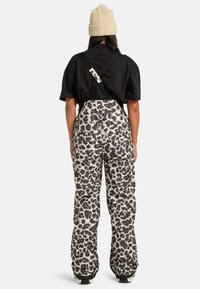 Osoba nosząca beżową dzianinową czapkę, czarną koszulkę z krótkim rękawem i białe spodnie śniegowe w czarne cętki z czarnymi szelkami, stojąca tyłem.