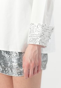 Bílá košile se stříbrnými flitry na manžetách. Flitry jsou hustě uspořádány, čímž vytvářejí texturovaný kontrast se hladkým materiálem.