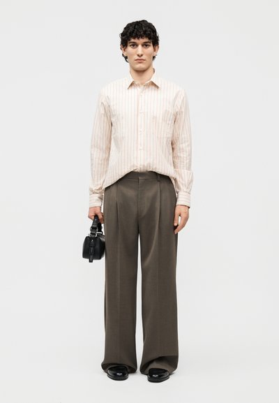 Junger Mann mit lockigem Haar, der ein cremefarben gestreiftes Hemd, weite braune Hosen und schwarze Schuhe trägt und eine kleine schwarze Handtasche hält.