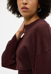 Suéter em borgonha com textura brilhante, gola alta e mangas largas. Material de malha macia apresenta um leve brilho e ajuste confortável.
