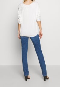 Eine Frau steht mit dem Rücken zur Kamera und trägt ein weißes Langarmoberteil, eine blaue enganliegende Jeans und schwarze High-Heels vor einem schlichten Hintergrund.