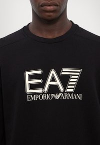 Juodas medvilninis džemperis su ryškiu baltu užrašu "EA7 EMPORIO ARMANI" su stilizuotomis raidėmis ir juostelėmis priekyje.