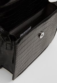 Borsa a mano in ecopelle nera con texture a coccodrillo, dotata di chiusura a scatto magnetico e una singola tasca interna. Etichetta del brand visibile all'interno.