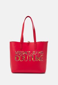 Czerwona torba na ramię wykonana z gładkiego materiału syntetycznego, z tekstem "VERSACE JEANS COUTURE" napisanym pogrubioną, wielokolorową czcionką.
