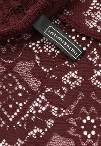 Bordeaux kantstof met bloemen- en geometrische patronen, voorzien van een zwart rechthoekig kledinglabel met "Intimissimi" in witte letters.