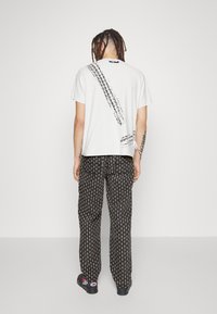 Persona con rastas de pie de espaldas, vistiendo una camiseta blanca con rayas diagonales negras y pantalones negros con estampado, además de zapatillas.