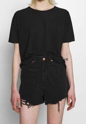 Person iført en sort tekstureret kortærmet skjorte, der er stoppet ned i højtaljede sorte denimshorts med frynset kant og kobberknapper.