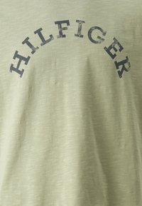 Lys grøn bomuld T-shirt med en blød tekstur, prydet med ordet "HILFIGER" i mørk marineblå, buet over brystområdet.