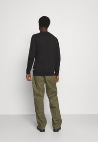 Svart långärmad skjorta med ribbade manschetter och nederkant, i kombination med olivgröna vida byxor. Slät textur, minimalistisk design och inga synliga mönster.