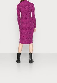 Robe ajustée à manches longues en rose vif avec un motif de vagues noires, arrivant au genou, fabriquée en matière extensible, associée à des bottines noires.