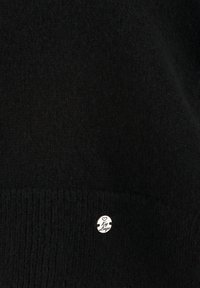 Schwarzer Strickpullover mit ribbeltem Saum und einem kleinen metallischen Logo-Tag mit dem Text "Key Lany" nahe dem unteren Ende. Weiche Textur.