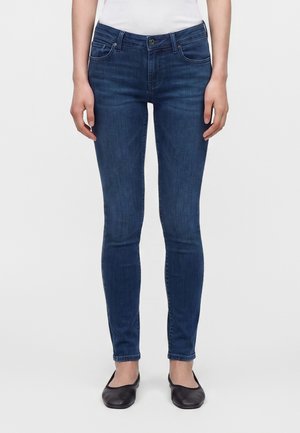 Jean slim bleu foncé porté avec un haut blanc et des ballerines noires sur une personne debout devant un fond clair uni.