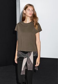 Oliwkowa bawełniana koszulka z luźnym krojem, okrągłym dekoltem i krótkimi rękawami. Noszona z czarnymi spodniami i swetrem w wzór argyle związanym w talii.