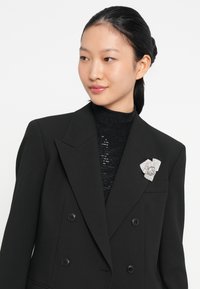 Blazer nero doppiopetto con un design strutturato, abbinato a un top nero testurizzato. Un fermaglio floreale in argento adorna il risvolto.