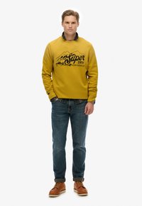 Sudadera amarilla con gráfico de montaña negra, superpuesta sobre una camisa de cuadros verdes. Combinada con jeans azules y botas marrones con cordones.