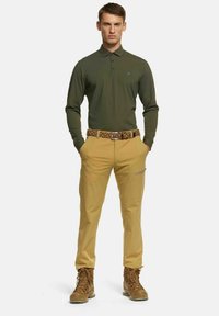 Grünes Langarm-Polohemd, beigefarbene Outdoor-Hosen mit Taschen, braune Wanderschuhe und ein gemusterter Gürtel. Schlichtes, funktionales Design.