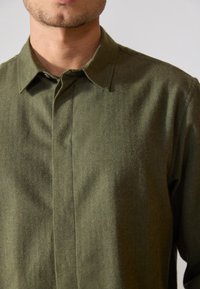 Camisa de botón de manga larga de color verde oliva, hecha de una suave tela texturizada; presenta un cuello puntiagudo y una costura vertical en el frente.