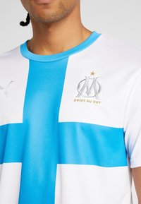 Valkoinen ja sininen urheiluhaalari, jossa on näkyvä sininen ristikkäinen kuvio, pyöreä kaulus, hopeinen logo sekä teksti "DROIT AU BUT".