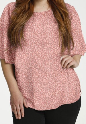 Woman with long brown hair wears a loose pink floral blouse with elbow-length sleeves and black pants, hand resting on hip.