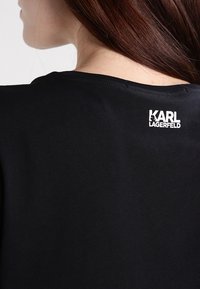 Mujer con cabello largo y castaño que lleva una camiseta negra con el logo blanco "KARL LAGERFELD" cerca del escote en la parte de atrás.