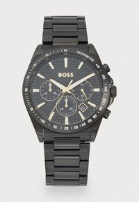 Montre en métal noir avec un cadran noir texturé, des accents dorés et trois sous-cadrans. Présente un bracelet en acier inoxydable et des repères de tachymètre.