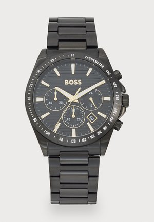Montre en métal noir avec un cadran noir texturé, des accents dorés et trois sous-cadrans. Présente un bracelet en acier inoxydable et des repères de tachymètre.