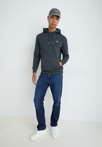 Donkergrijze hoodie met een kangoeroezak, verstelbare koordjes, gedragen met donkerblauwe jeans en witte sneakers, onder een grijze pet.