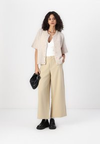 Cardigan corto a maniche corte beige chiaro con bottoni, abbinato a pantaloni a gamba larga color crema. Una borsa nera e scarpe chunky nere completano l'outfit.