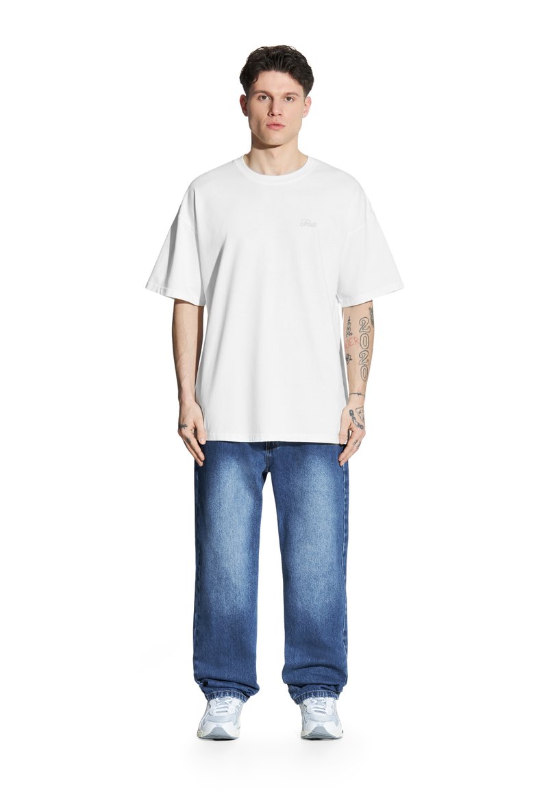 Biała, oversize'owa bawełniana koszulka z krótkim rękawem, zestawiona z niebieskimi dżinsami. Model nosi szare i białe trampki. Prosty detal z logo na koszulce.