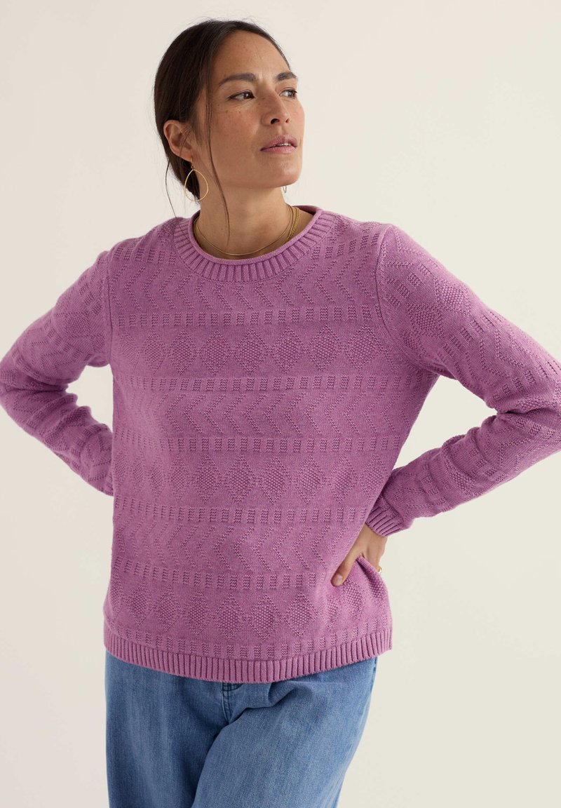 Naine, kellel on seljas tekstuuriga lilla kampsun geomeetriliste mustritega ja sinised teksad, seisab käed puusadel, vaadates küljele.