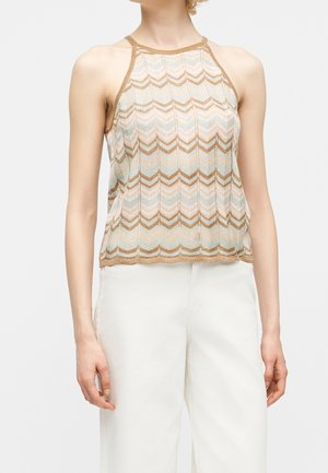 Femme portant un haut sans manches beige et blanc à motif zigzag, associé à un pantalon blanc taille haute sur un fond uni.