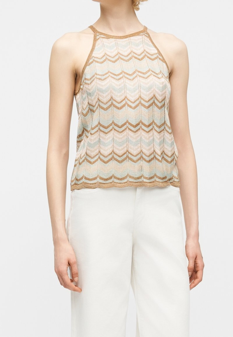 Femme portant un haut sans manches beige et blanc à motif zigzag, associé à un pantalon blanc taille haute sur un fond uni.