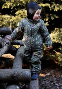 Laps tikitud, lillemustrilise rohelise jope ja sobivate pükstega, musta mütsiga, logidel looduslikus keskkonnas seismas.