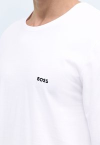 Valkoinen pitkähihainen T-paita, joka on valmistettu puuvillasta ja jossa on musta brodeerattu "BOSS"-logo vasemmalla rintataskualueella. Sileä rakenne, pyöreä kaula-aukko.