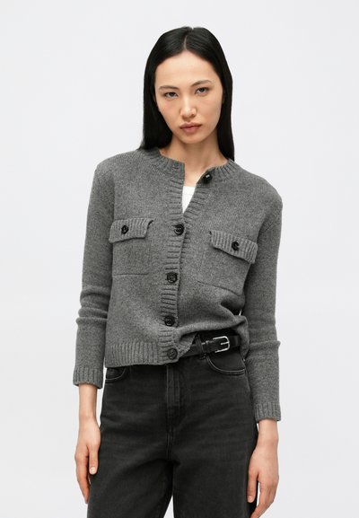 Femme portant un cardigan gris boutonné avec des poches à rabat et un jean noir taille haute avec une ceinture noire sur un fond uni.