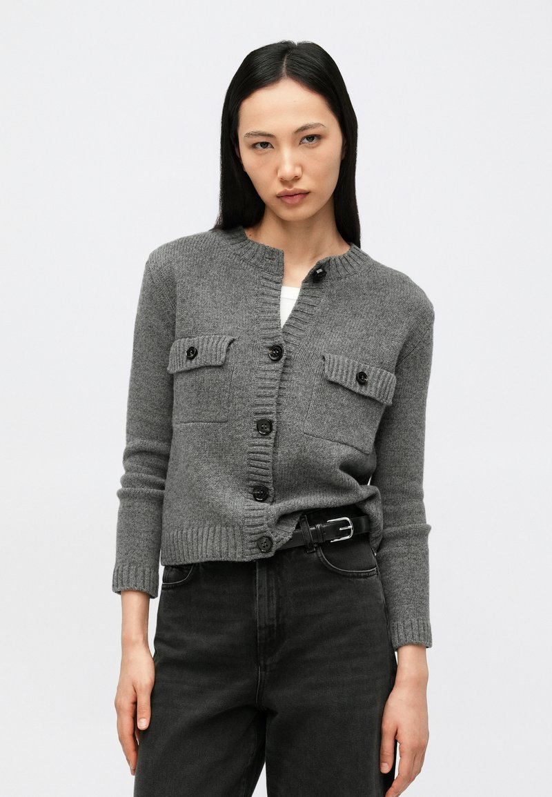 Γυναίκα που φοράει γκρι ζακέτα με κουμπιά και τσέπες με καπάκια, καθώς και μαύρο ψηλόμεσο τζιν με μαύρη ζώνη, μπροστά σε απλό φόντο.