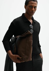 Bruine canvas tas met een gestructureerde afwerking, zwarte trui en grijze accessoire eroverheen gelegd; ronde zonnebril ligt op de kraag van de trui.