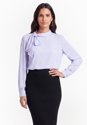 Frau trägt eine hellviolette Bluse mit einer Halschleife und einem hoch taillierten schwarzen, gerippten Rock und posiert mit einer Hand auf der Hüfte.