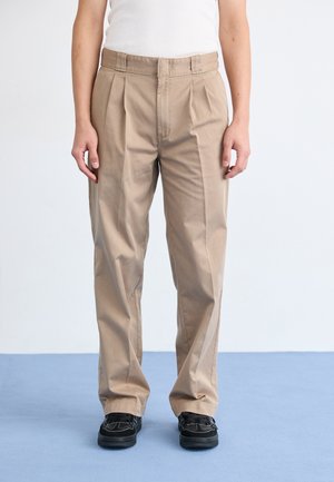 Beige plooibroek met een rechte pijp, gemaakt van katoen. De tailleband is glad en de stof heeft een zachte textuur. Gemaakt in combinatie met zwarte schoenen.